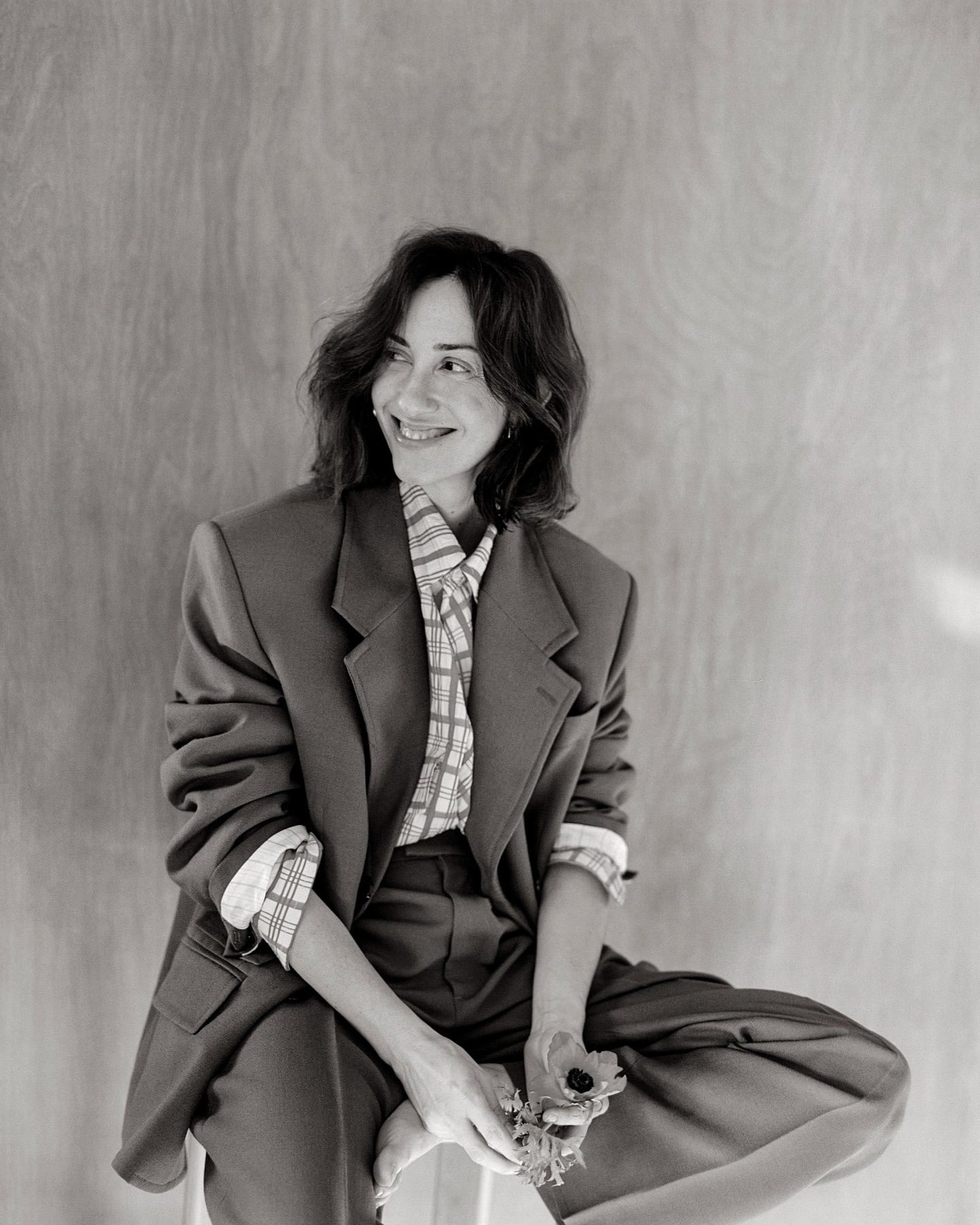 Black and white portrait of an adult woman smiling and wearing a blazer and patterned shirt, seated in front of a wood wall.