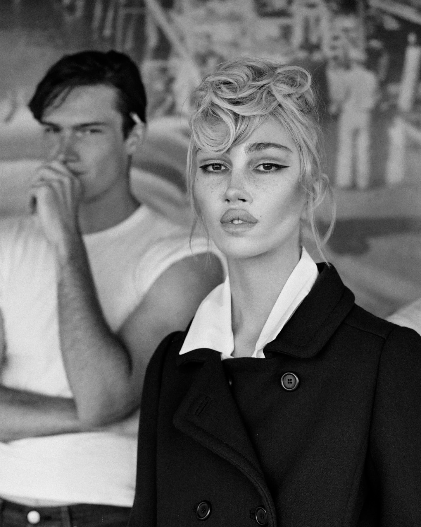 Black and white portrait of a blonde woman in a coat with a man in the background, in front of a faded cityscape.