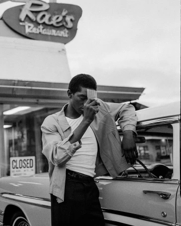 Young man stands by a classic car near Rae's Restaurant, holding comb to his face in a monochrome, vintage-style photograph.