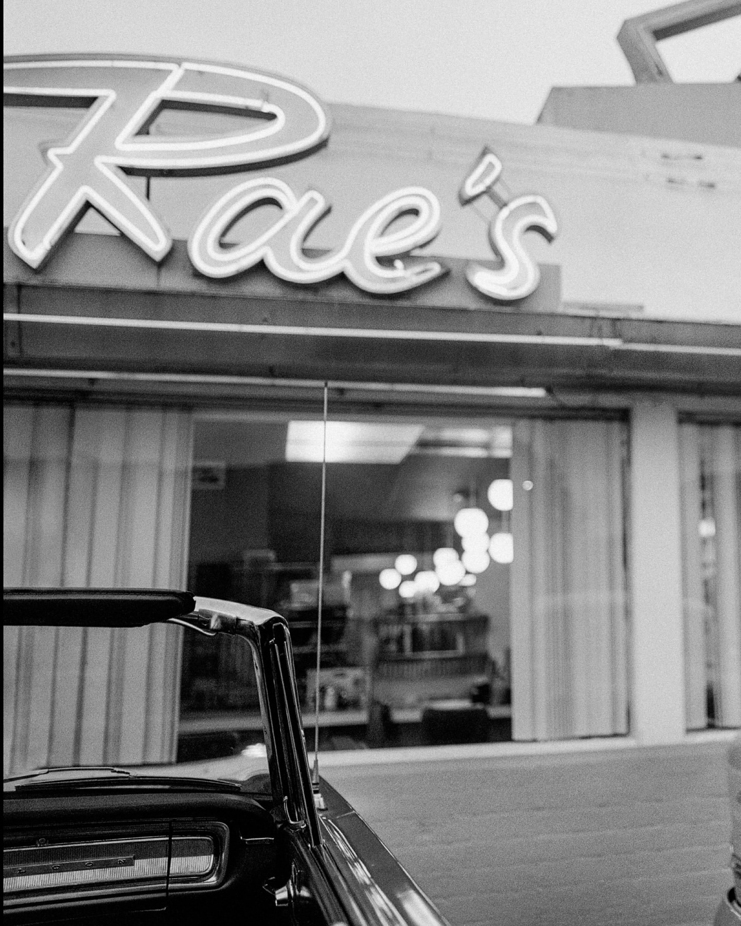 Exterior of Rae's diner, black and white photograph with neon sign, classic car parked in front, retro architectural style.