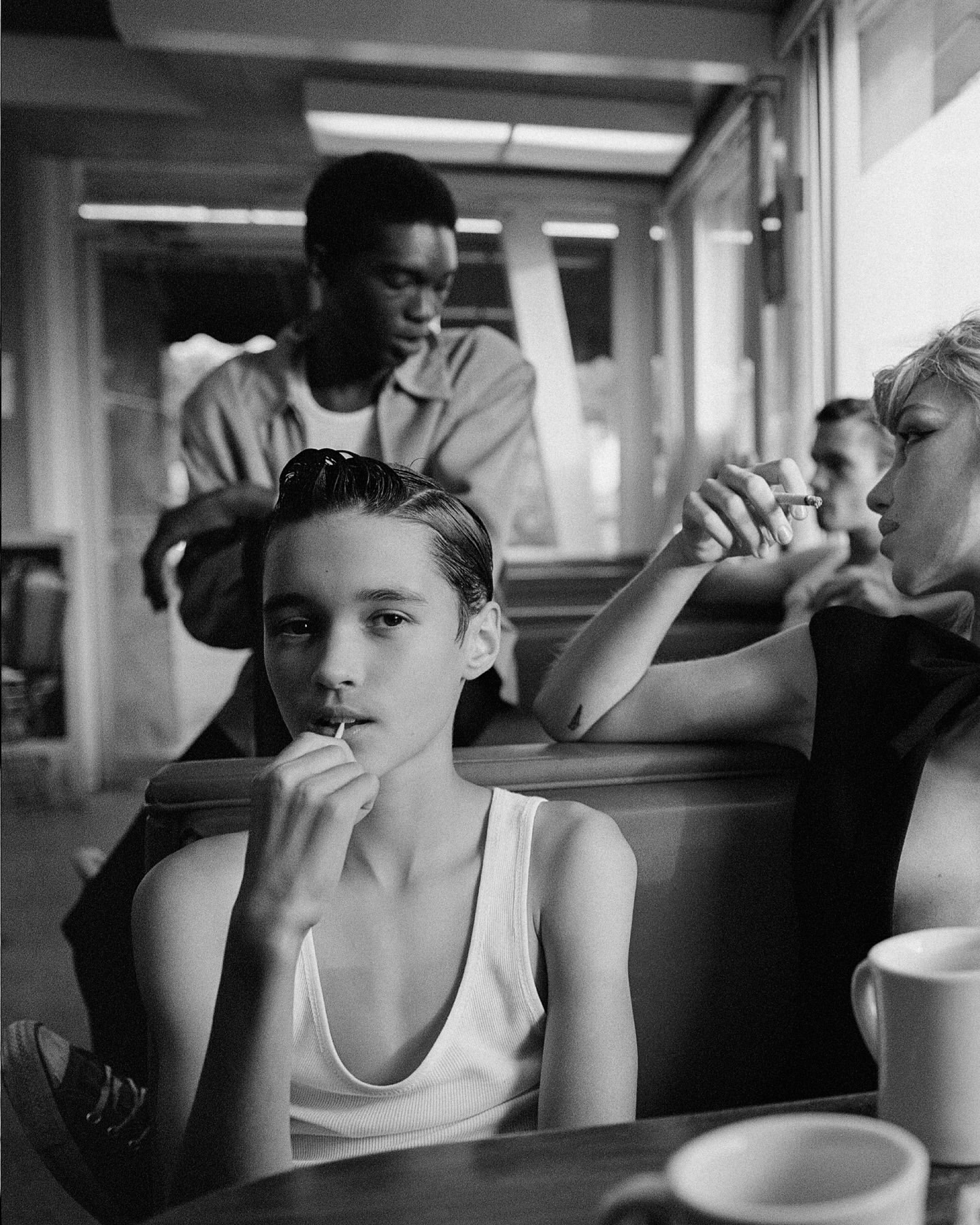 Monochrome photo of teenagers in a diner booth. One boy looks directly at the camera while others smoke and sit around him.