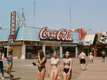 Several people in swimwear walking along a boardwalk past food stalls with large Coca-Cola signage.