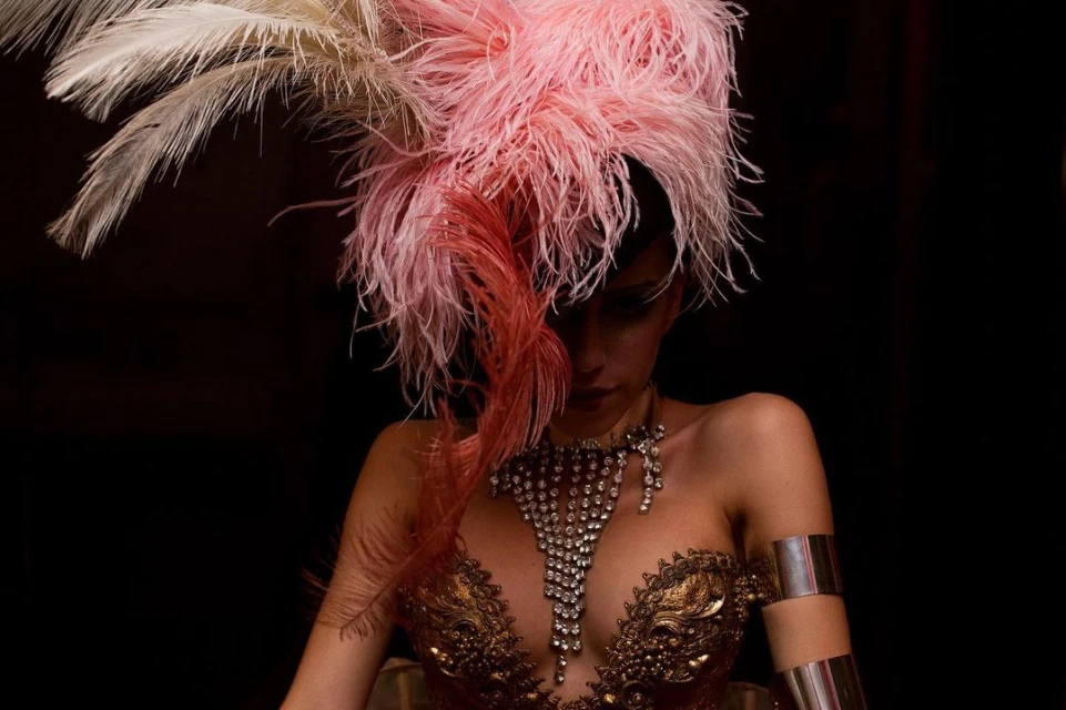Portrait of a woman with an ornate gold dress, silver arm cuffs, and a large feather headpiece against a dark background.
