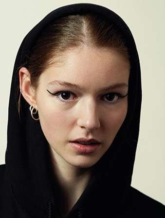 Close-up portrait of a young adult woman with fair skin, wearing a black hooded top and hoop earrings, against a neutral background.