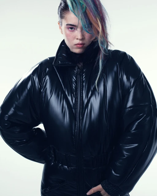 Studio shot of a young woman with dyed hair wearing a shiny black puffer jacket, posing against a white backdrop with strong lighting.