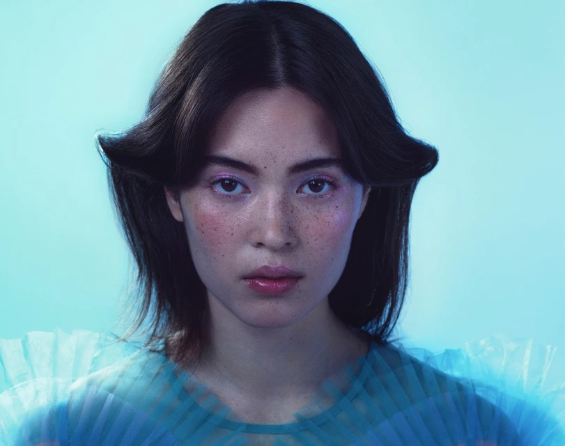 Close-up portrait of an adult female with dark hair and pale skin, wearing a light blue sheer top against a light blue background.