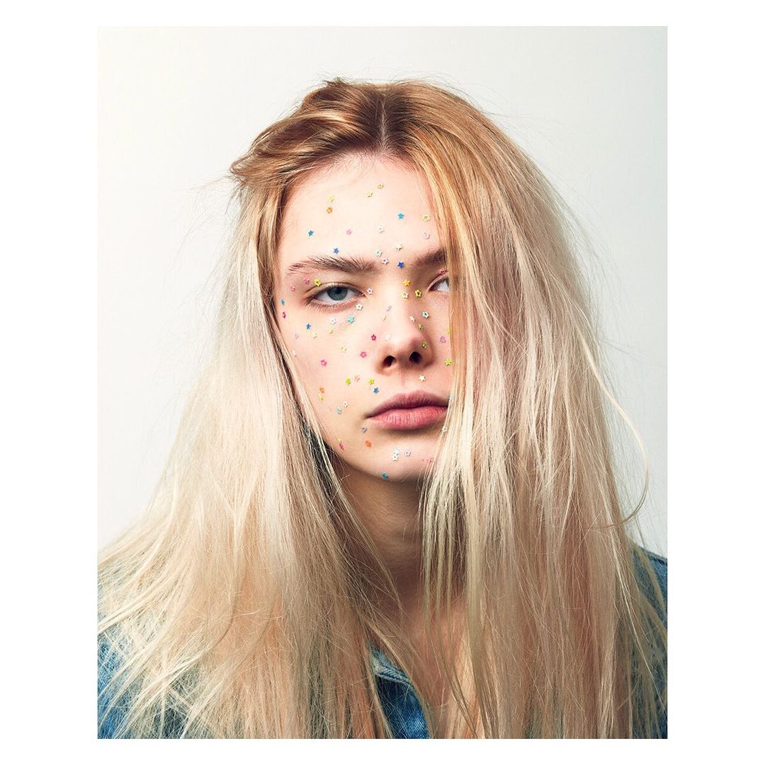 Close-up portrait of a young adult woman with blonde hair and colorful gems on her face, against a light background.