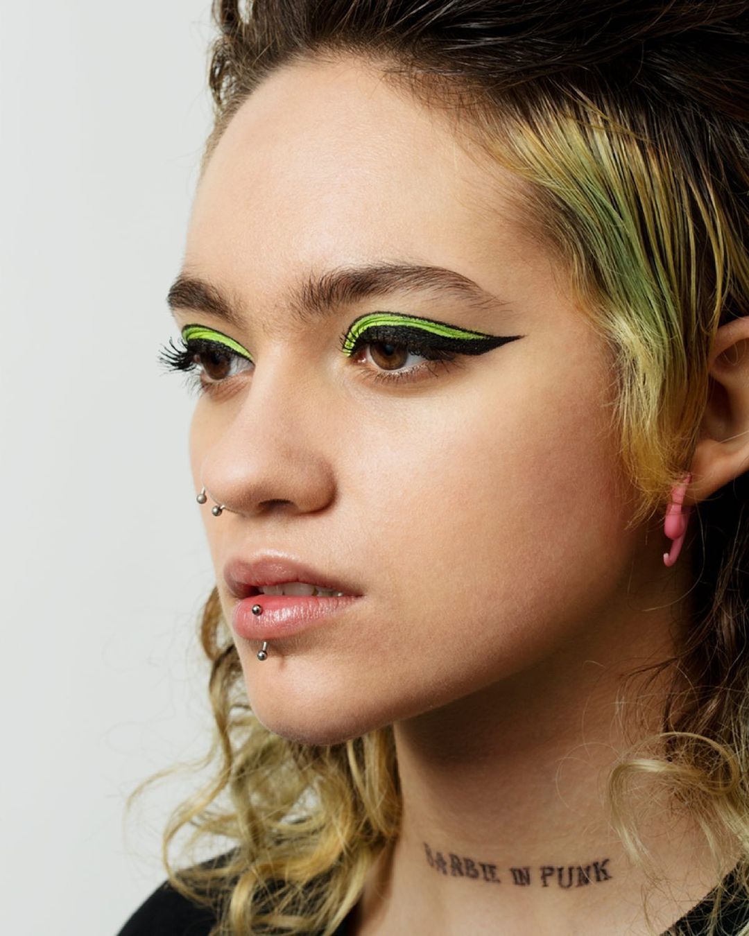 Close-up studio shot of a young adult woman with dark hair, bright green winged eyeliner, pink earrings, and a visible tattoo.