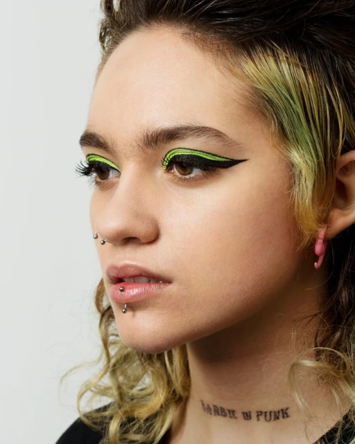Close-up studio shot of a young adult woman with dark hair, bright green winged eyeliner, pink earrings, and a visible tattoo.