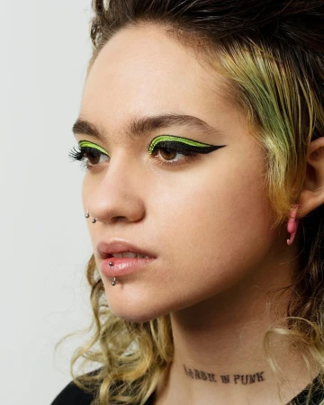 Close-up studio shot of a young adult woman with dark hair, bright green winged eyeliner, pink earrings, and a visible tattoo.