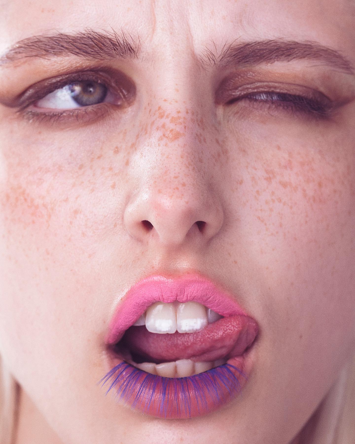 Close-up beauty shot of a young adult woman with pink lipstick and purple eyelash extensions winking with her tongue sticking out.