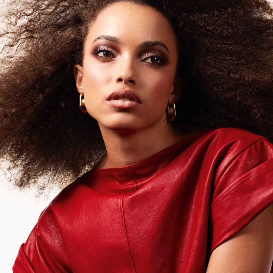 Portrait of a woman with curly hair wearing a red top and gold hoop earrings against an off-white backdrop in a photo studio.