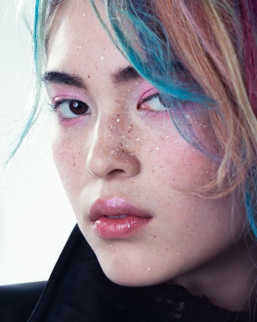 Close-up portrait of a young woman with rainbow-colored hair and pink makeup, featuring a light skin tone and subtle freckles.