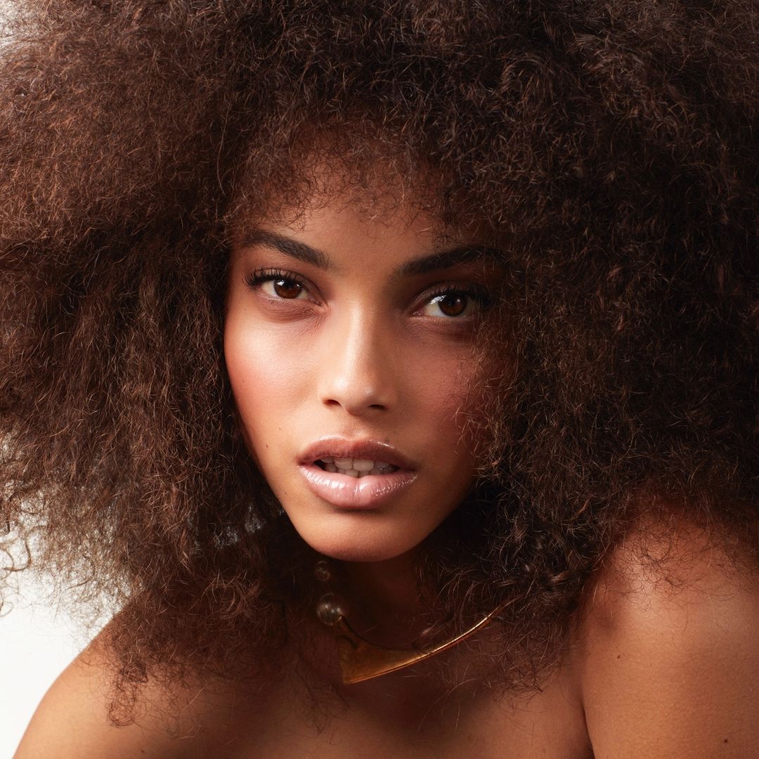 Portrait of a woman with brown curly hair, wearing a gold collar necklace, and a neutral facial expression, in a studio setting.
