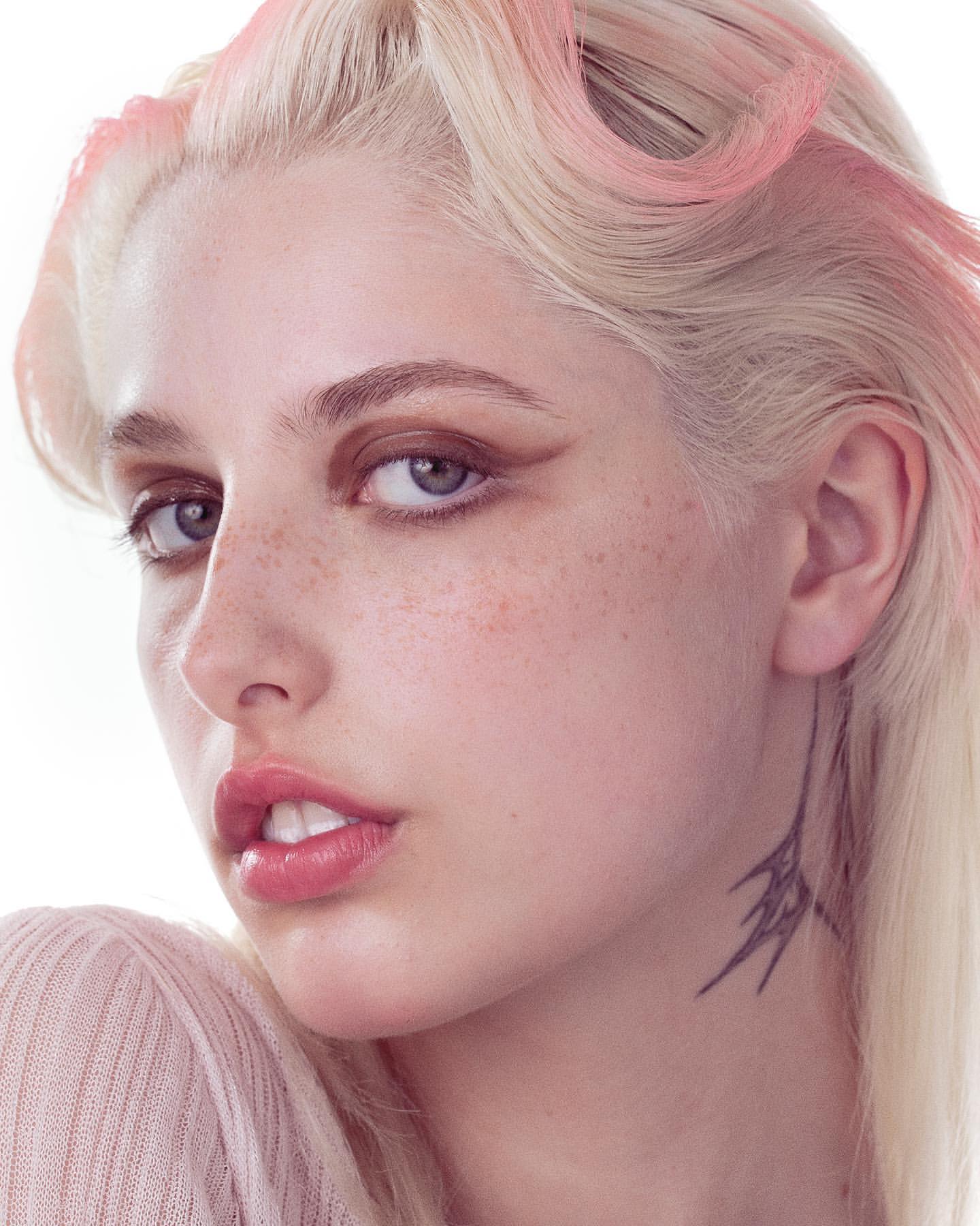 Close-up studio portrait of an adult female with fair skin, light blonde hair with light pink highlights, and a delicate neck tattoo.