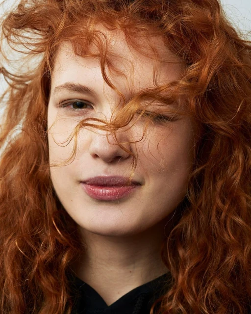 Close-up portrait of a smiling, young adult female with curly red hair against a neutral background, face partially obscured by her hair.
