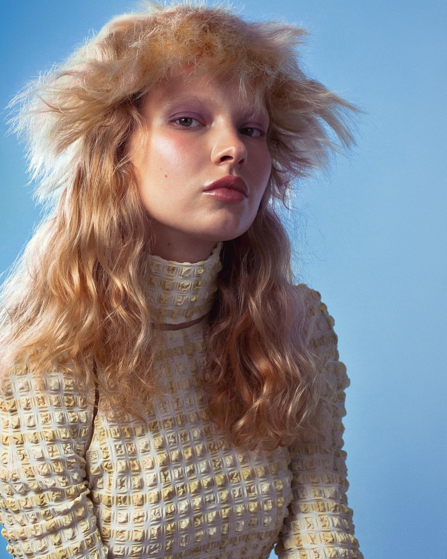Portrait of a woman with wavy blonde hair and a high-necked textured beige top against a pale blue backdrop in a beauty fashion style.
