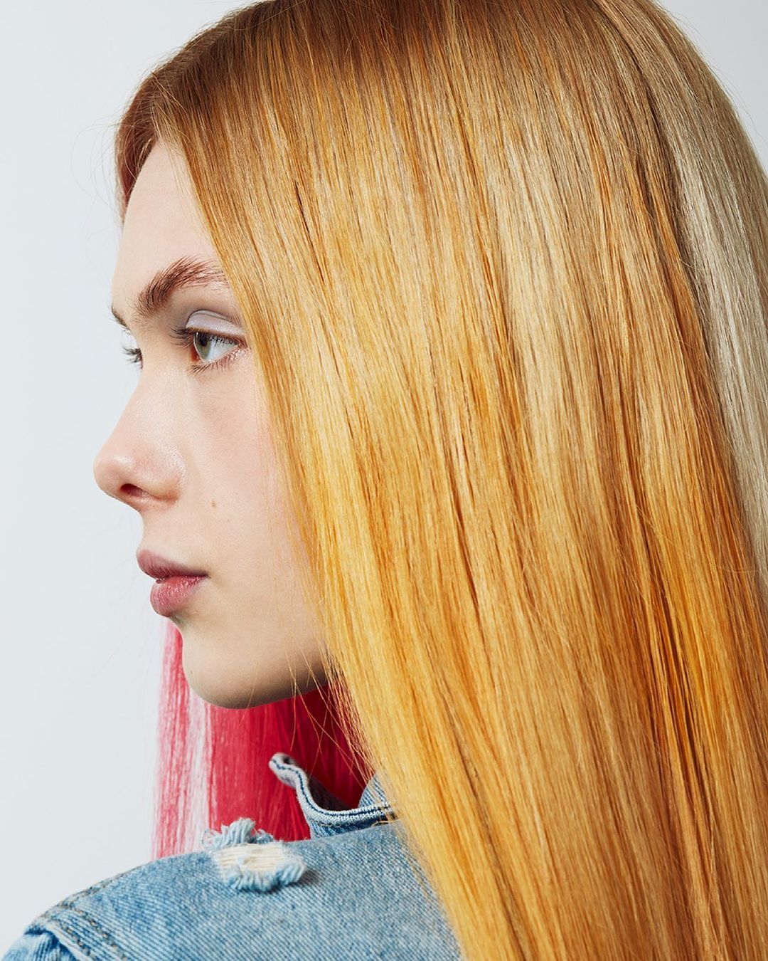 Profile portrait of a blonde woman with brightly colored hair highlights wearing a denim jacket in front of a white background.