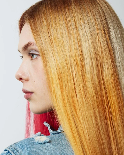 Profile portrait of a blonde woman with brightly colored hair highlights wearing a denim jacket in front of a white background.