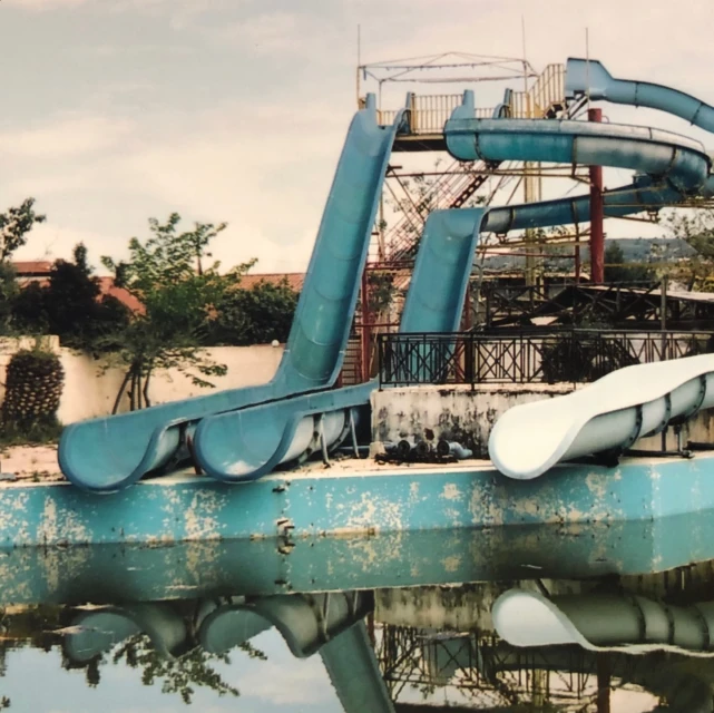 Overgrown abandoned water park featuring blue and white water slides, empty swimming pool, and decaying structures against a cloudy sky.