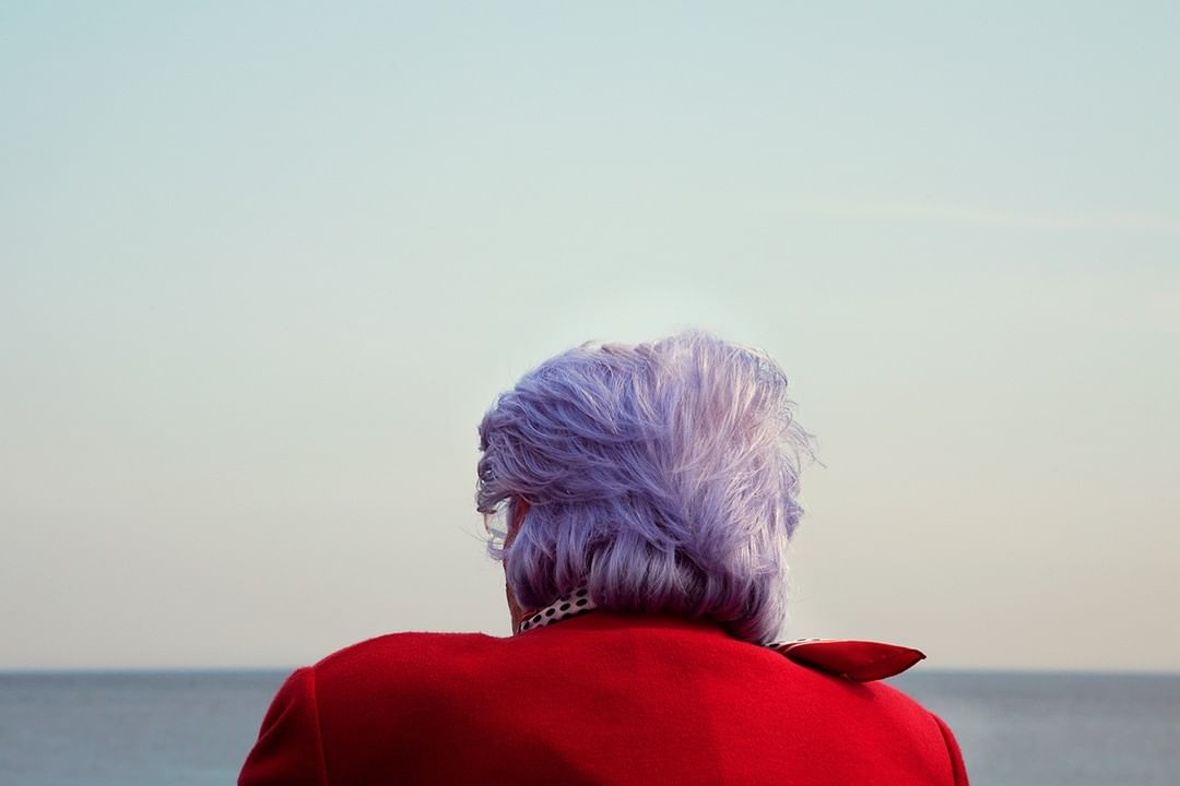 Back view of elderly woman with lavender hair in a red jacket, gazing at the ocean under a clear sky, enjoying peaceful outdoor scene.