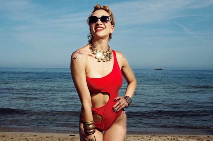 Woman in red swimsuit smiles on beach with ocean and blue sky background, accessorized with necklaces, bracelets, and sunglasses.