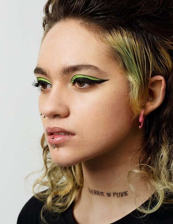 Close-up of a young adult female with green graphic eyeliner, multiple piercings, and a neck tattoo, showcasing a beauty and alternative fashion aesthetic.