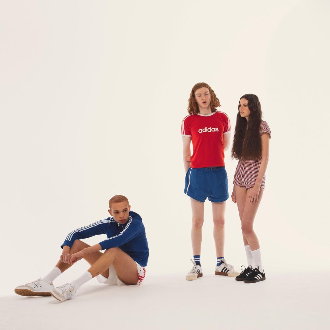 Three fashion models in 70's style athletic wear pose in a bright studio. Two are standing and one is sitting.