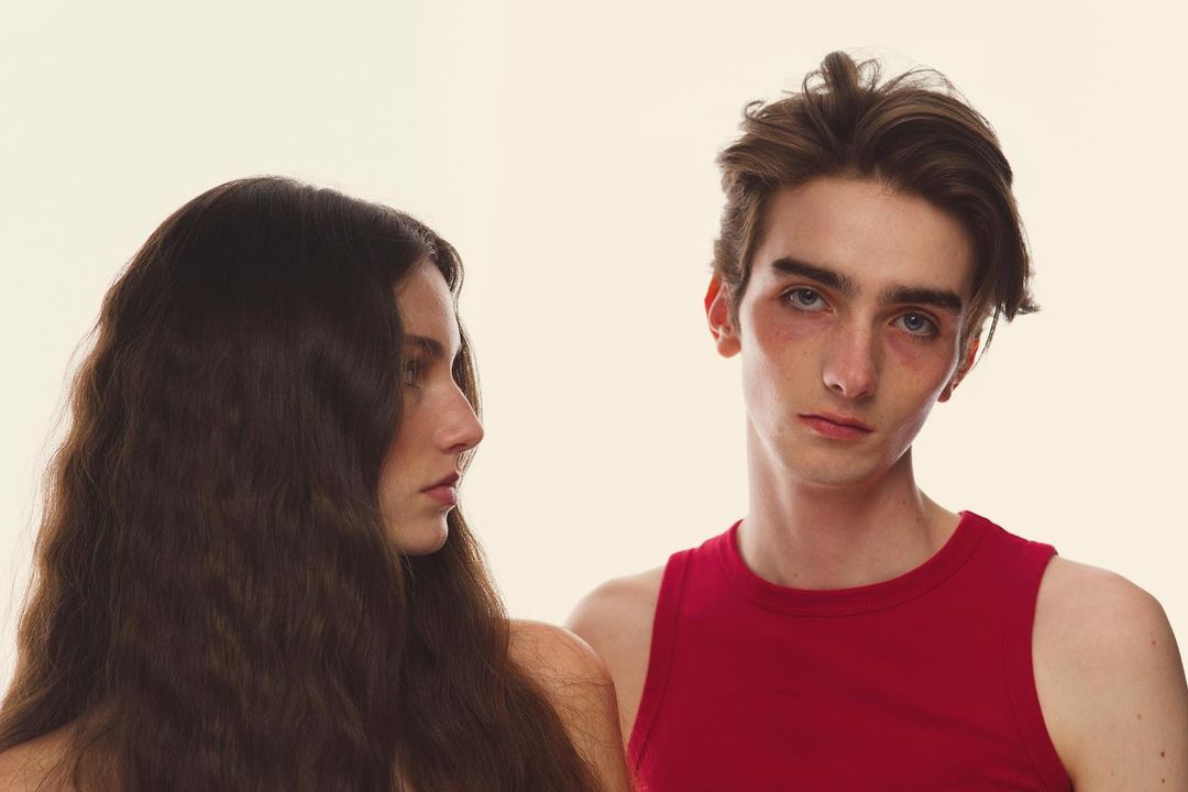 Fashion headshot of a woman in profile with long brown hair and a man in a red tank top posing against a white backdrop.