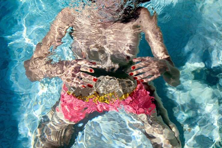 Underwater view of a person swimming in a pool with pink floral trunks and red nails, reflecting light patterns on the water surface.
