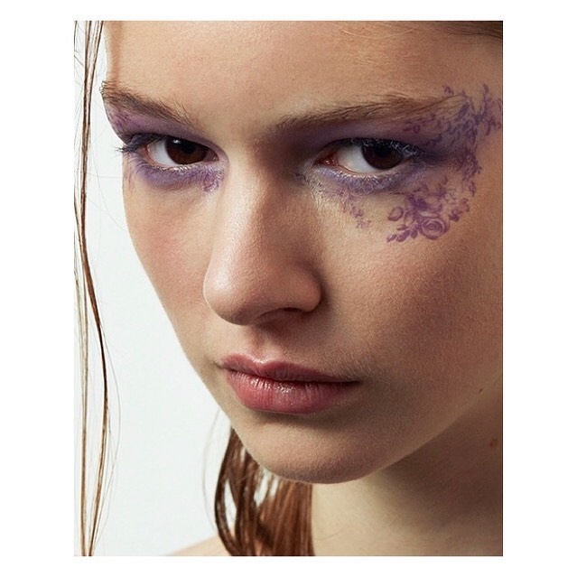 Close-up of a young adult female with artistic purple floral eye makeup in a vertical studio shot against a bright white background.