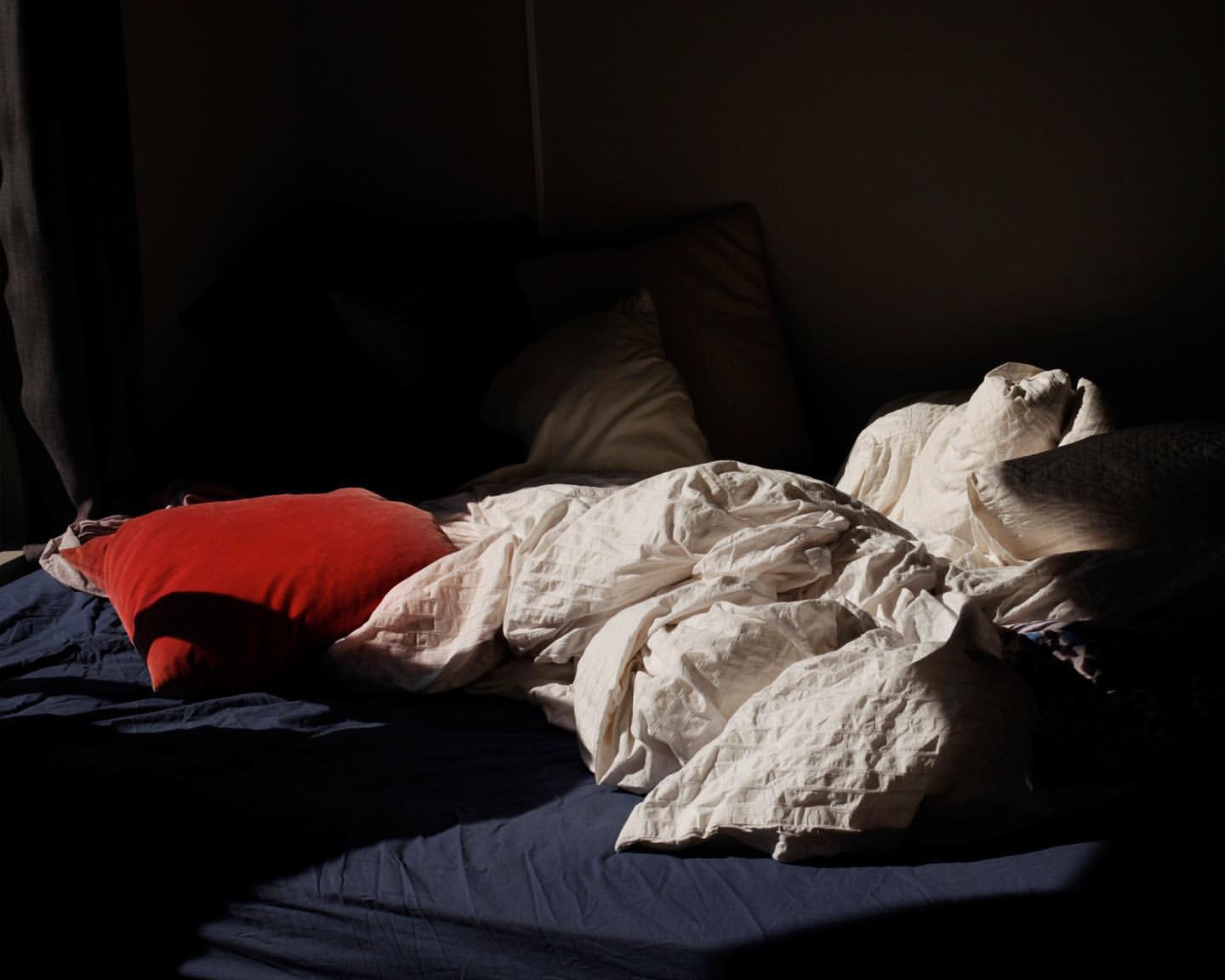 Bed with blue sheets and wrinkled white comforter, red pillow, soft lighting, creating a cozy and inviting atmosphere.