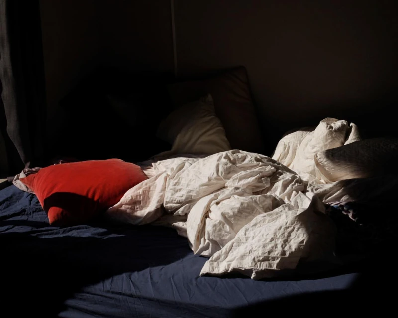 Bed with blue sheets and wrinkled white comforter, red pillow, soft lighting, creating a cozy and inviting atmosphere.