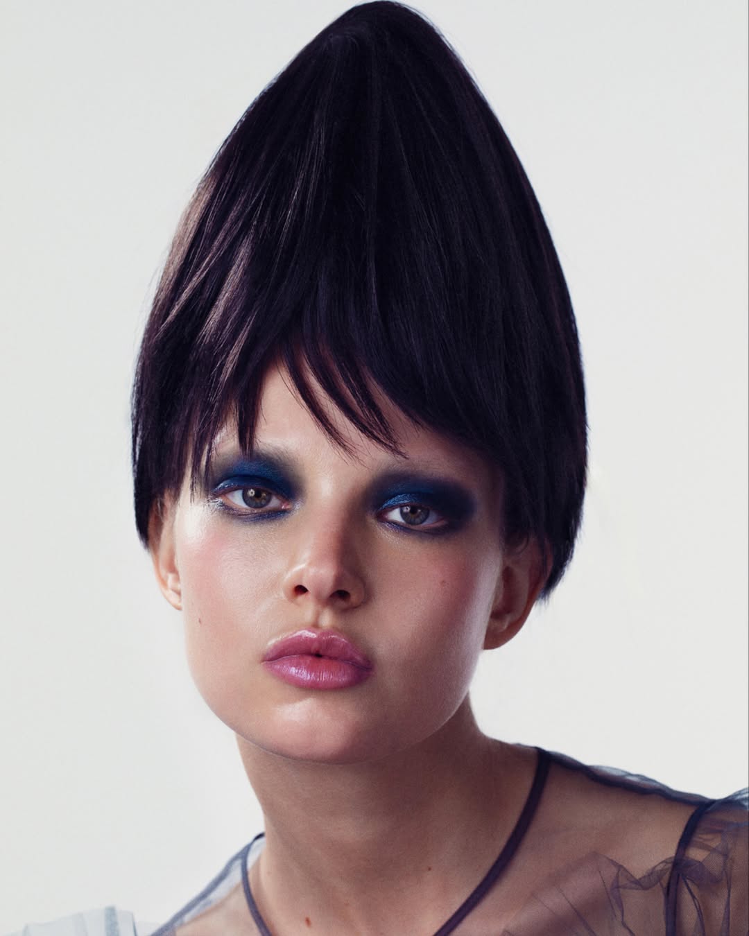 Close-up portrait of a woman with dramatic blue eyeshadow and an avant-garde dark hairstyle, set against a plain white background.
