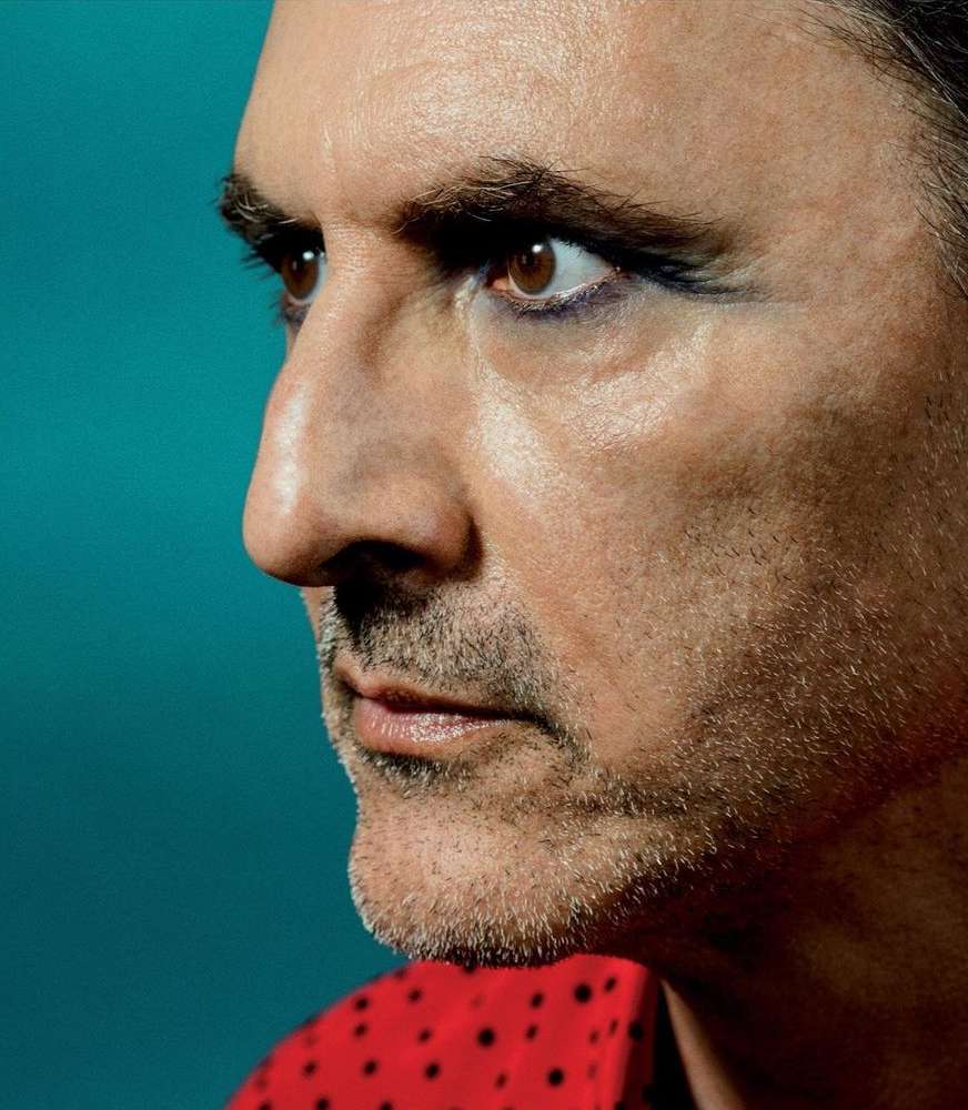 Close-up profile of a man with fair skin, stubble, and makeup, wearing a polka dot shirt against a turquoise background.