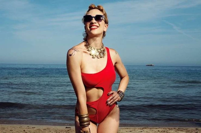 Smiling woman wearing a red one-piece swimsuit and sunglasses at the beach by Chloe Claverie