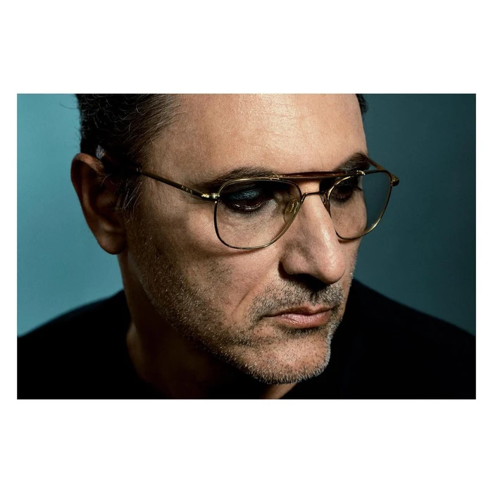 Close-up portrait of a man wearing thin-framed aviator glasses and dark clothing against a teal backdrop