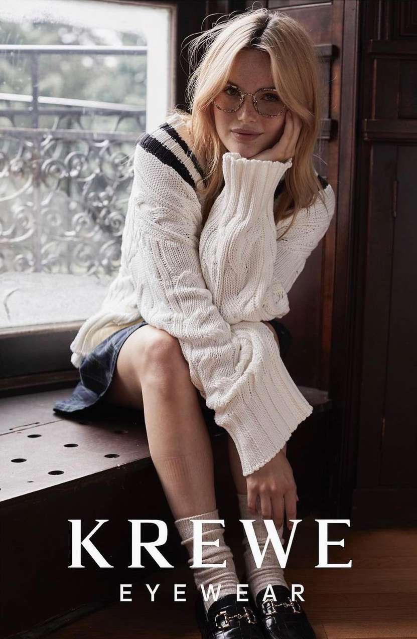 Blonde woman wearing glasses and a cream-colored cable knit sweater poses seated near a window, accessorized with black penny loafers and socks.