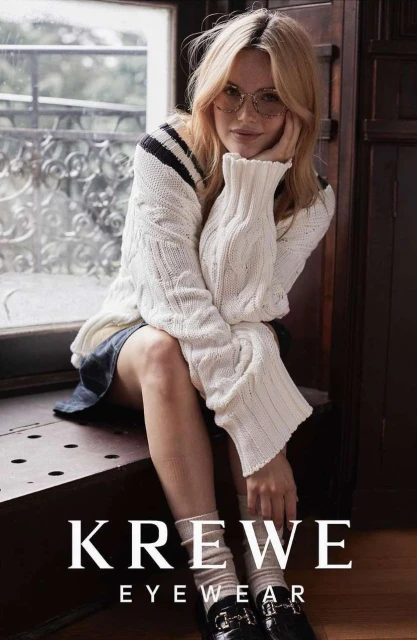 Blonde woman wearing glasses and a cream-colored cable knit sweater poses seated near a window, accessorized with black penny loafers and socks.