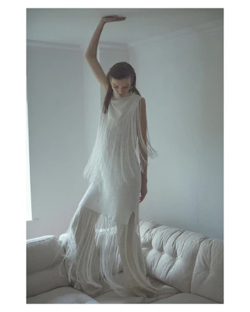 A fair-skinned woman in a sleeveless, floor-length fringe dress stands on a tufted sofa in an indoor setting near a window.