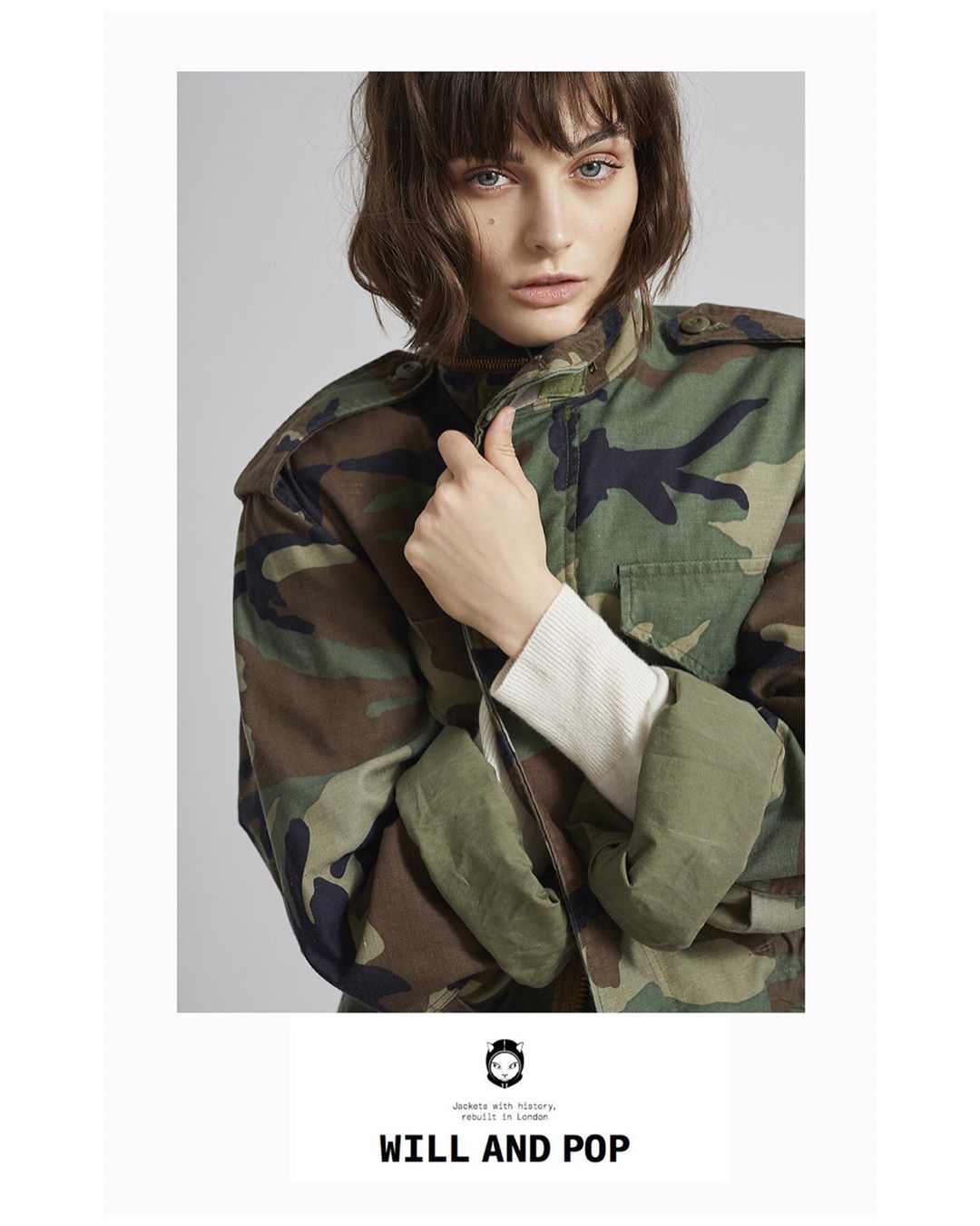 Portrait of a young adult female model in a camo jacket, with neutral facial expression and shoulder-length brown hair, posing indoors.
