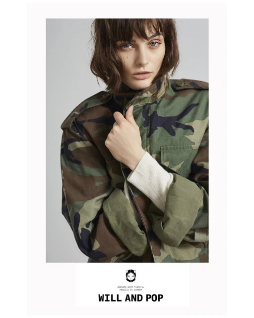 Portrait of a young adult female model in a camo jacket, with neutral facial expression and shoulder-length brown hair, posing indoors.