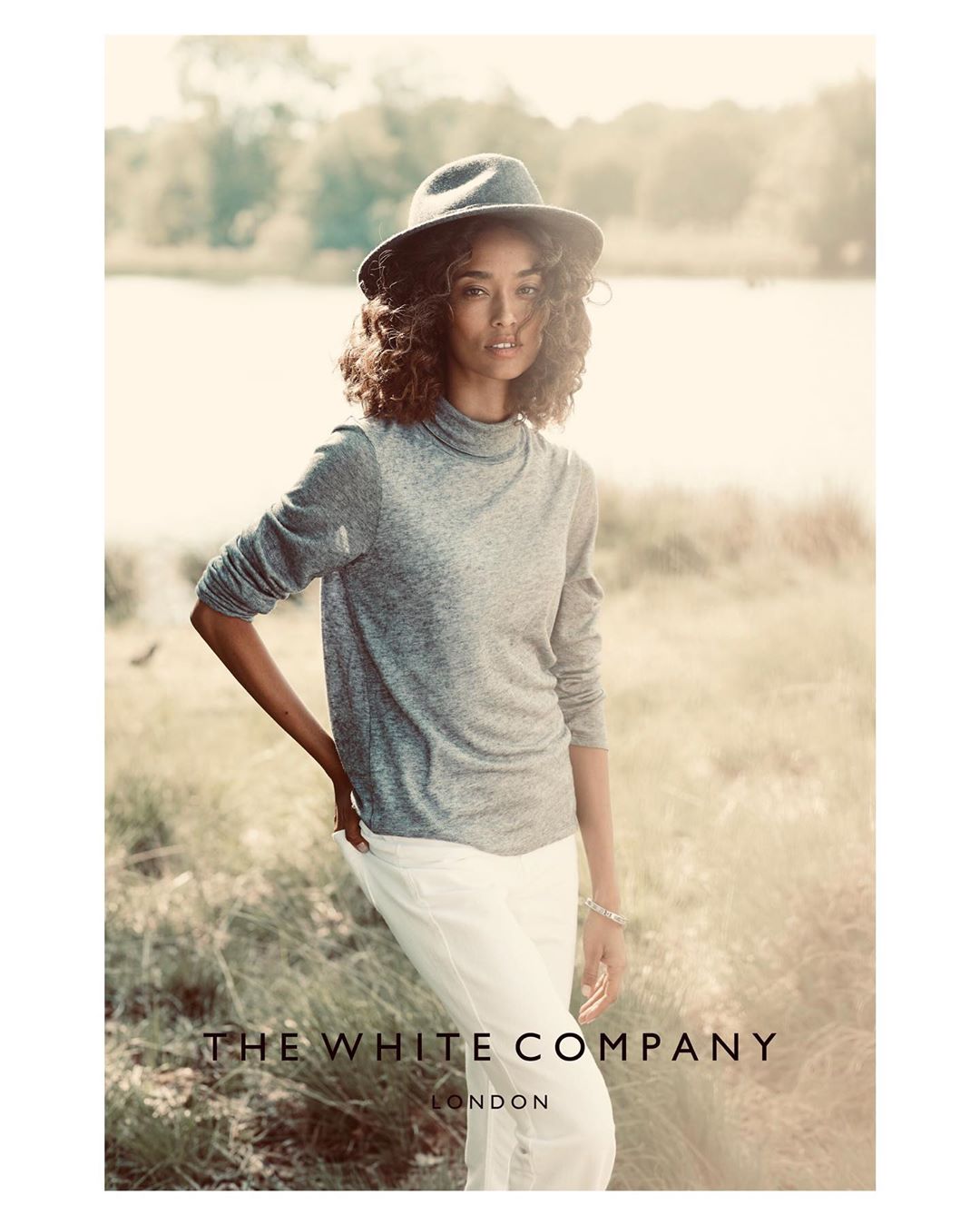 An adult female model poses in a gray long-sleeve shirt and white pants, wearing a dark hat in an outdoor field setting.