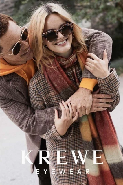 Smiling woman in sunglasses with man embracing her in autumnal outerwear and scarf, posing in an outdoor setting. Eyewear ad for Krewe.