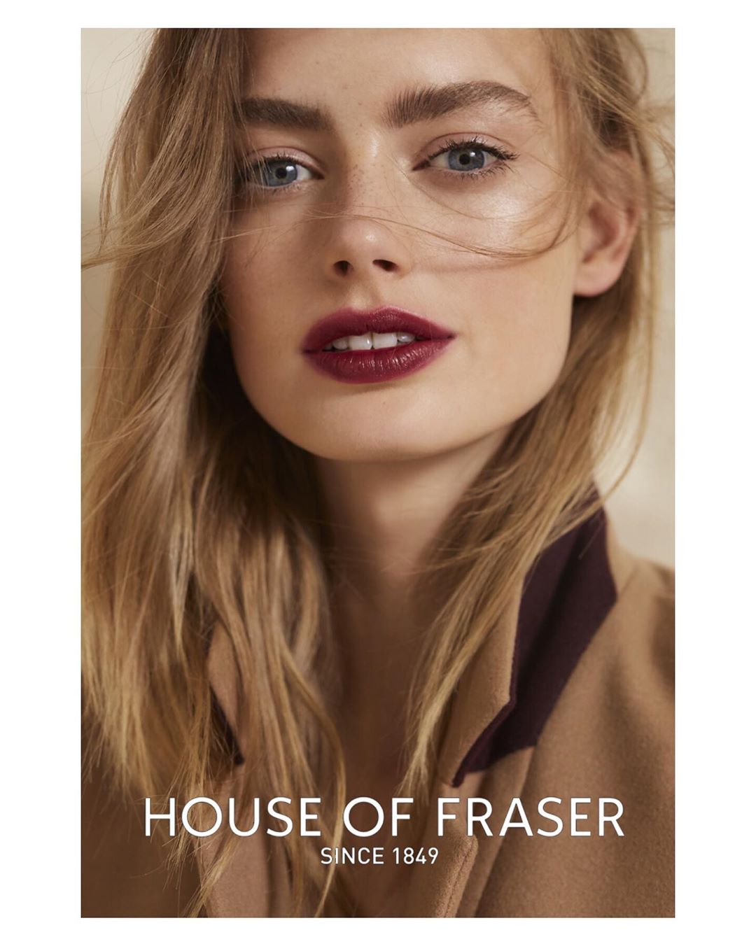 Close-up portrait of a blonde female model with striking features and dark red lipstick, wearing a tan jacket, in a studio setting.