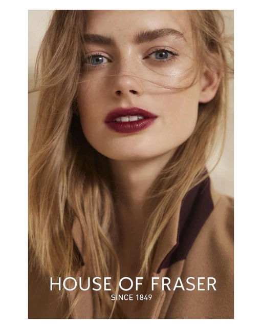 Close-up portrait of a blonde female model with striking features and dark red lipstick, wearing a tan jacket, in a studio setting.