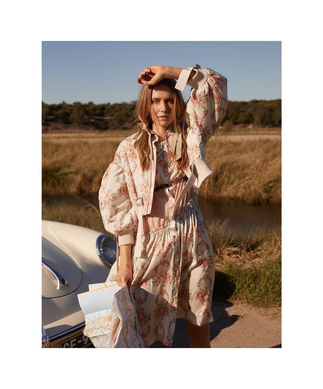 Woman in floral jacket and dress poses next to a classic car in a field holding a map in the daytime, fashion editorial shot.