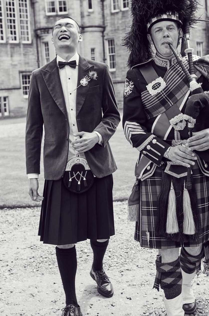 Black and white image of an Asian man in a kilt smiling next to a bagpiper in traditional garb outside a stone building.