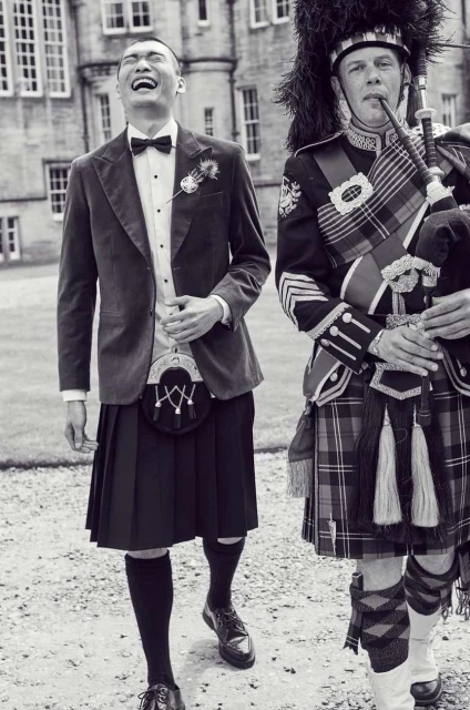 Black and white image of an Asian man in a kilt smiling next to a bagpiper in traditional garb outside a stone building.