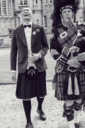Black and white image of an Asian man in a kilt smiling next to a bagpiper in traditional garb outside a stone building.